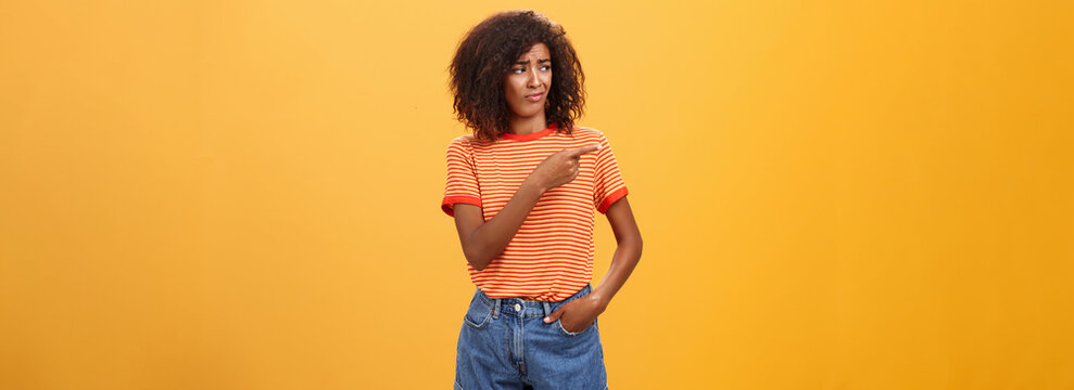 Girl looking at doubtful unimpressive perfomance of model. Portrait of displeased confused good-looking dark-skinned female with afro hairstyle looking and pointing left with scorn and indifference