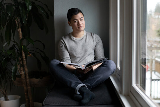 Asian American trans man sits by the windowsill.