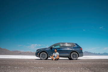 Fototapeta na wymiar Man sitting by car at Bonneville salt flat with blue sky in background. Male traveler is relaxing at tranquil landscape during sunny day. He is enjoying vacation at famous place.