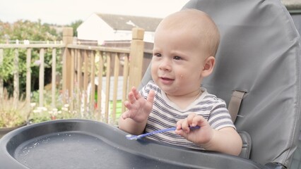 Woman feeding Tired child with spoon. Mom feed crying Sad baby Boy with pureed food. Mom feeding kid in baby chair outside in the garden . Summer time. Toddler nutrition