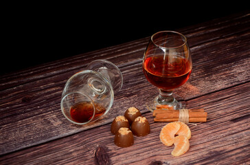 Two glasses of cognac, cinnamon sticks, tangerine slices and chocolates on a wooden table.