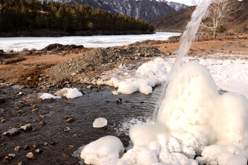 A stream of a small mountain stream flows down onto an ice block of frozen water on the bank of a beautiful river on a winter day.
