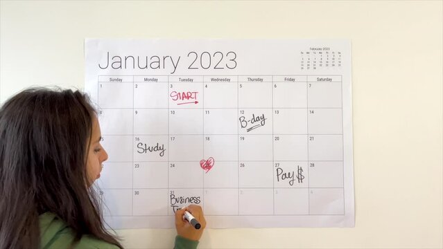 Woman planning a new month by writing bussiness travel on a 2023 wall calendar in black marker