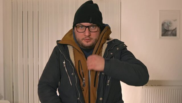 ironic caucasian male wearing and zipping multiple layers of winter jacket facing cold season at home with higher price cost of gas and living