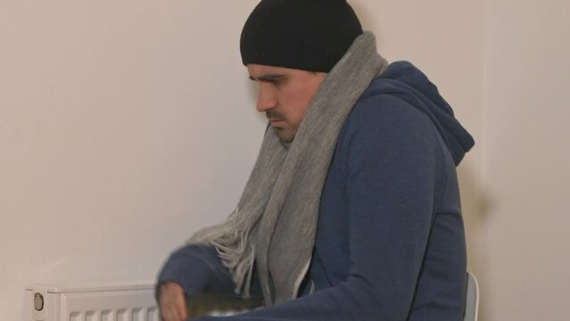 Freezing young man with hoodie, scarf and hat sitting next to a heater and shivering with cold, energy crisis