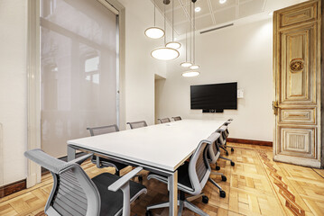 Boardroom of a stately vintage building with a long white wooden table with gray swivel chairs and hardwood flooring and designer lamps