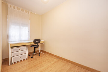 A mostly empty room with a white study table with a chest of drawers and a black study swivel chair with poor back support