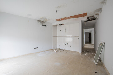 An unfinished empty room with kitchen tiles on one wall and heavy damage caused by water leaks