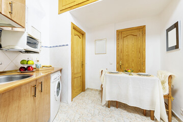Old kitchen with a similar wooden worktop, wooden cabinets and individual appliances of the same color and a dining area with a tablecloth and the table mounted in one corner