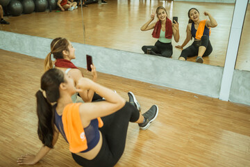 two asian women sitting in front of mirror taking selfie together using mobile phone post workout...