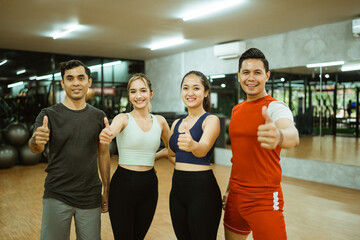 Fototapeta na wymiar Group of diverse young friends in sportswear standing with thumbs up during break in gym