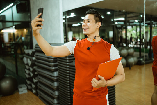 male instructor standing holding clipboard making video call with smartphone after workout in gym