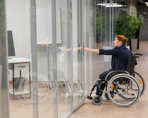 Red-haired caucasian woman in a wheelchair trying to open the door in the office. 