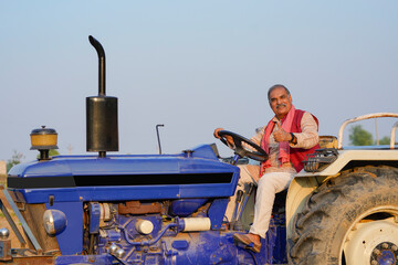 Indian farmer sitting on tractor and showing thumps up at agriculture field.