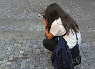A young long-haired tourist girl takes pictures with a DSLR camera, squatting down (her face is...