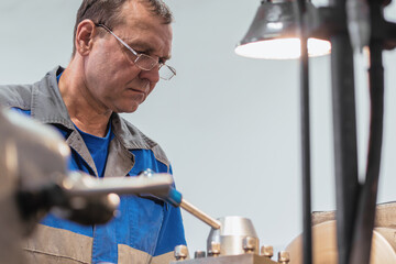 Portrait of turner with glasses 50-55 years old at work. Elderly metal turner works in workshop on machine behind job..
