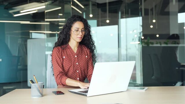 Curly brunette employee works in a modern office at workplace at a laptop computer focused and confident woman programmer web developer writing code typing programming business female worker in casual