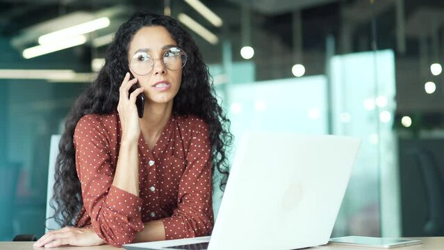 confident busy attractive business woman employee talking on the cell phone sitting in modern office at the computer desk at workplace indoors curly dark haired brunette female speaks conversation
