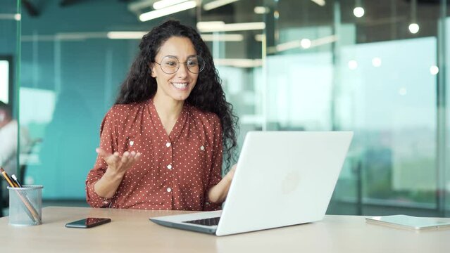 Happy young beautiful business woman with curly hair and glasses Hispanic positive employee talking on video call using laptop for remote communication and meeting conference in a modern office