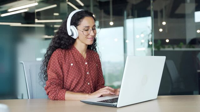 Curly hispanic brunette young woman employee working on laptop computer and listening music sounds or radio in headphones in modern office Pleasant girl student worker enjoying at workplace indoors
