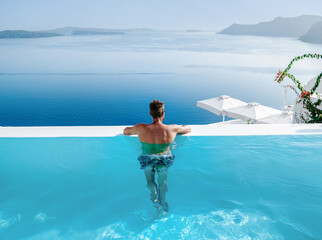 Santorini Greece Oia, young men in swim shorts relaxing in the pool looking out over the caldera of...