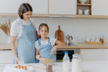 Smiling mother and daughter prepare tasty cookies, girl whisks ingredients in bowl with beater, helps mum at kitchen, prepare festive dinner, modern home interior. Family, cooking, lifestyle concept