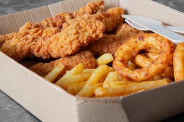 Breaded fried chicken breast with fries and onion rings, in a cardboard tray with wooden cutlery. Environmentally friendly disposable plastic free container
