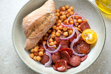Beige plate with fried chorizo sausage, chickpeas, red onion and crusty bread, middle close-up, elevated view, horizontal shot