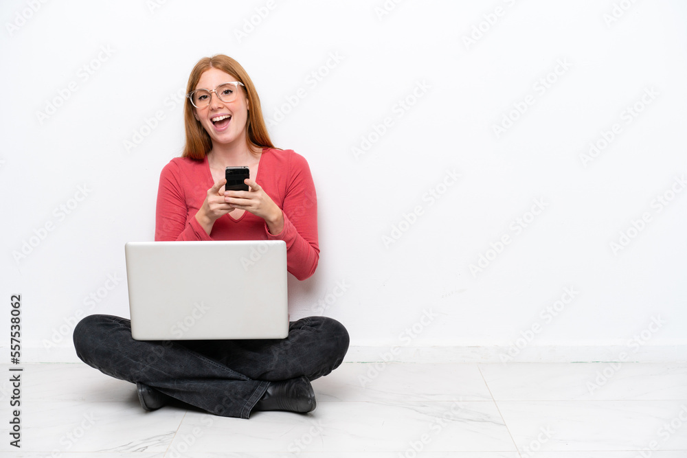 Poster Young redhead woman with a laptop sitting on the floor isolated on white background surprised and sending a message