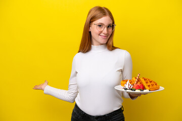Young redhead woman holding waffles isolated on yellow background extending hands to the side for inviting to come