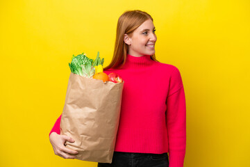 Young redhead woman holding a grocery shopping bag isolated on yellow background looking side