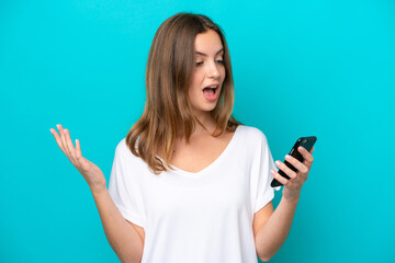 Fototapeta premium Young caucasian woman isolated on blue background looking at the camera while using the mobile with surprised expression