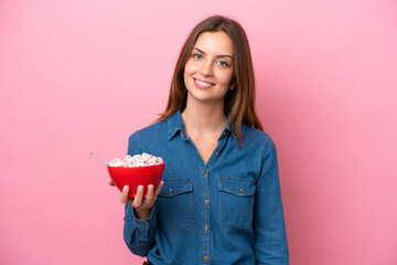 Young caucasian woman holding a bowl of cereals isolated on pink background smiling a lot