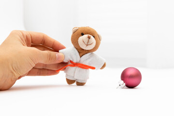 The hand holds a teddy bear in a white kimono with an orange belt. Nearby lies a Christmas ball