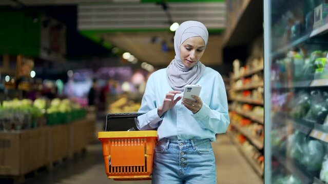 Young muslim woman in hijab Buyer customer checking shopping list using smartphone, purchasing in grocery store in supermarket female shopper pick goods use mobile phone in hypermarket or food market