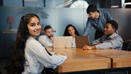 Indian ethnic businesswoman looking at camera posing in office on blurred background of brainstorming diverse office workers multiethnic multiracial coworkers discussing work with laptops at table