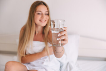 Happy beautiful woman drink water during wake up on morning at the bedroom. Smiling girl holding transparent glass on bed.