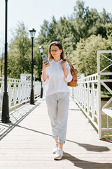 Satisfied female student using smartphone with bag over her shoulder while walking in park. Smiling stylish young woman wearing casual clothes chatting, browsing internet on phone outdoors