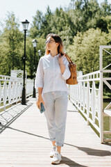 Satisfied carefree female student wears glasses and throws bag over her shoulder walking in park on sunny day, looking away