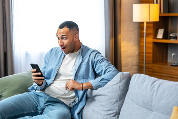 Surprised happy african man holding phone looking at cellphone read good news in sms sit on sofa,...