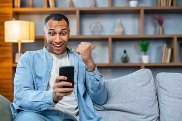 Man at home on sofa using smartphone, celebrating victory in living room reading happy news...