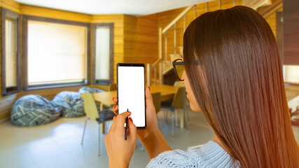 Woman is using phone. Girl with smartphone with her back to camera. Phone with white screen in hands of girl. Lady with telephone in wooden house. Concept app for interior design. Art focus