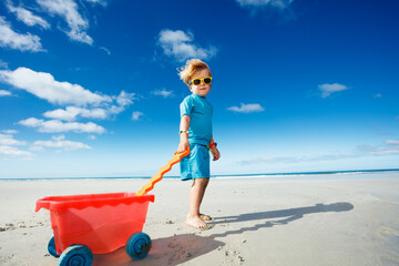 Happy blond boy play with toy cart on white sand ocean beach