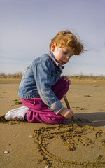 Bambina bionda che scrive sulla sabbia del bagnasciuga con i capelli al vento