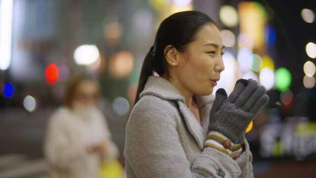 Attractive Asian woman feeling cold and wearing knit gloves while enjoy travel and shopping at Shibuya, Tokyo city, Japan in winter night with crowd of people walking backgrounds on holiday vacation.