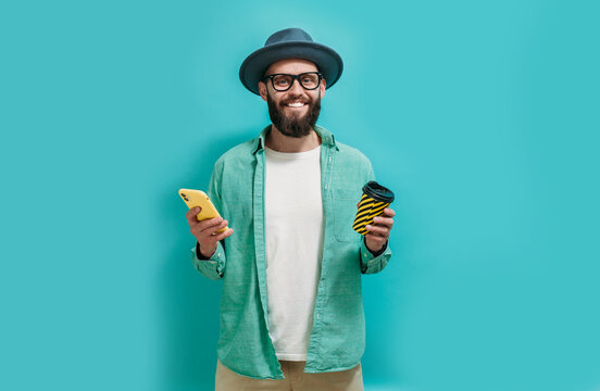 A Shot Of A Smiling Hipster Guy With A Thick Beard Holding A Mobile Phone, Drinking Coffee To Go, Dressed In Casual Clothes, A Hat And Glasses, Reading A Message Over Blue Studio Background