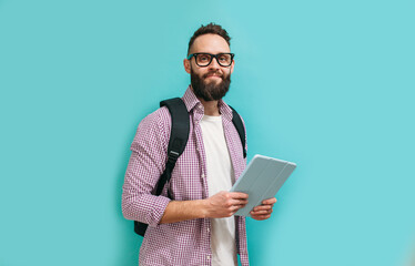 Portrait of handsome smiling student in casual wear with backpack while holding digital tablet over blue studio background