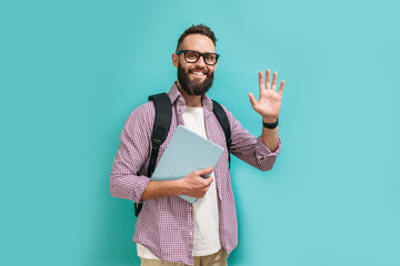 Portrait of handsome smiling student in casual wear with backpack while holding digital tablet over blue studio background