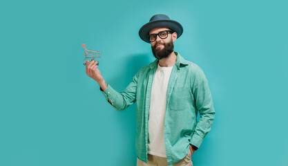A young hipster man dressed casual holding a mini shopping cart isolated on blue studio background....