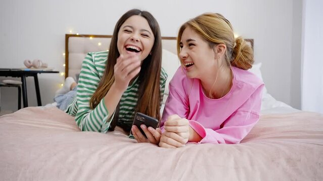 Cute teens friends girls lie on bed, chat online on smartphone, share photos. Friendships concepts